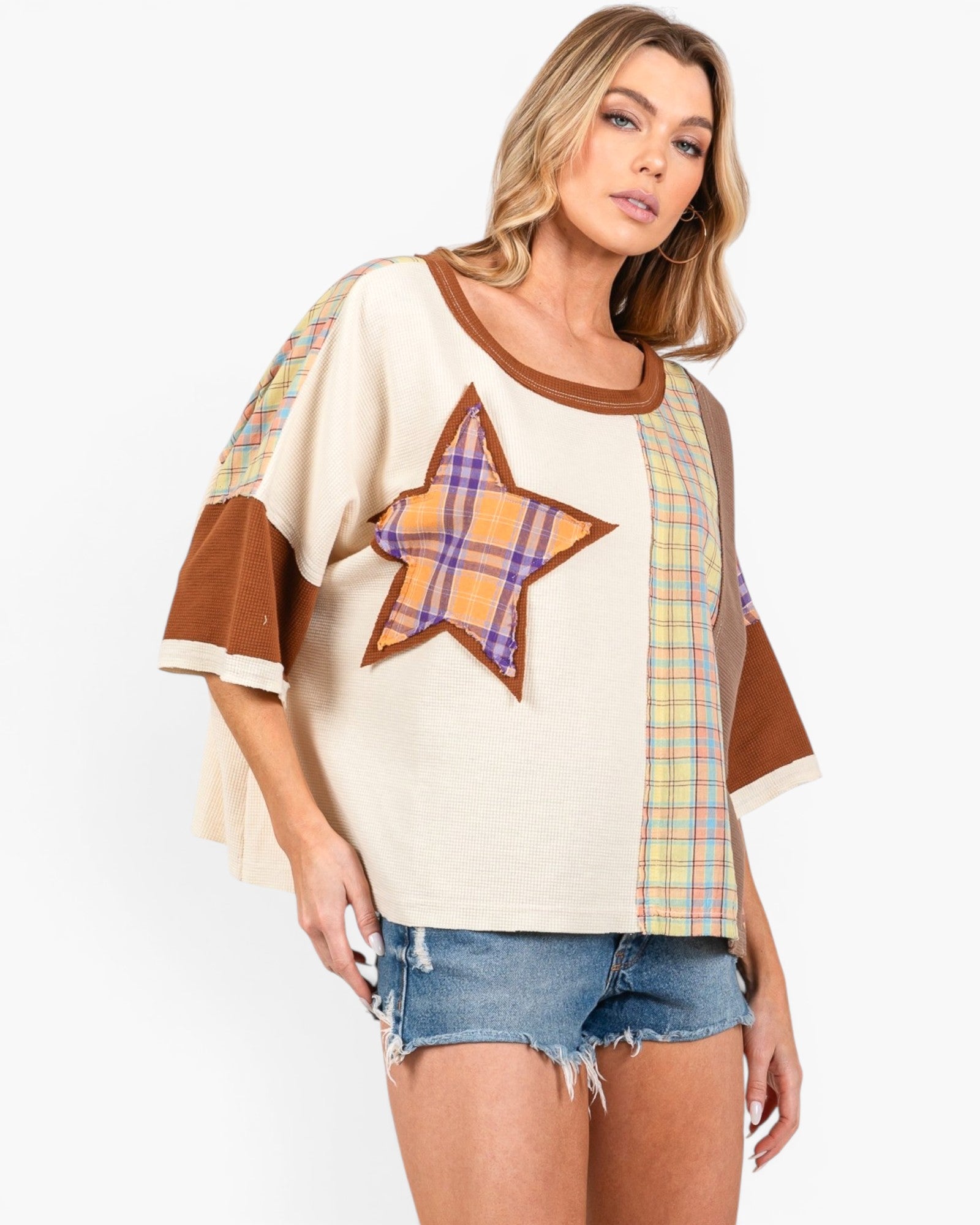 A woman modeling a cream-colored patchwork top with brown trim, a large plaid star on the chest, and plaid sleeve panels, paired with denim shorts against a light gray background.