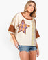 A woman modeling a cream-colored patchwork top with brown trim, a large plaid star on the chest, and plaid sleeve panels, paired with denim shorts against a light gray background.