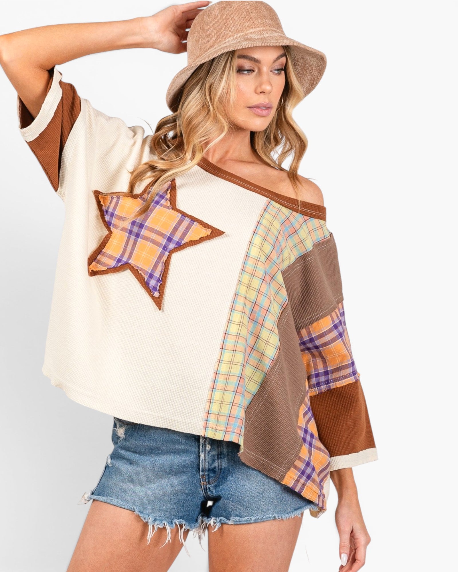 A woman modeling a bohemian-style off-the-shoulder patchwork top with a star patch, plaid details, and brown trim, paired with denim shorts and a beige bucket hat against a light gray background.