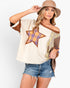 A woman modeling a cream-colored off-the-shoulder patchwork top with a large plaid star on the chest, brown trim, and denim shorts, wearing a tan hat against a light gray background.