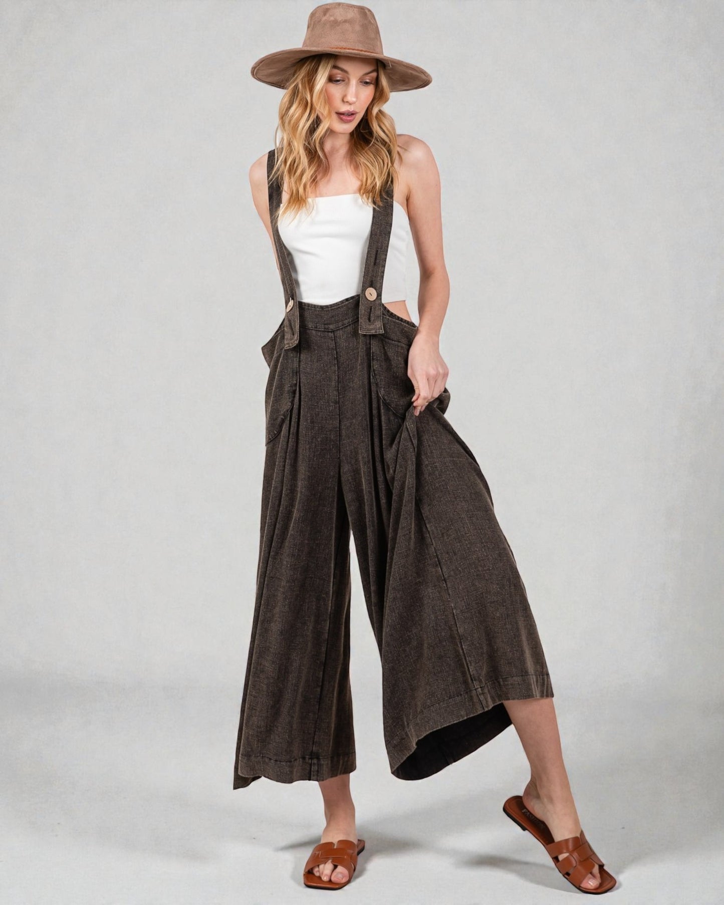 Woman wearing brown wide-leg pants with suspenders and a white top on a plain background