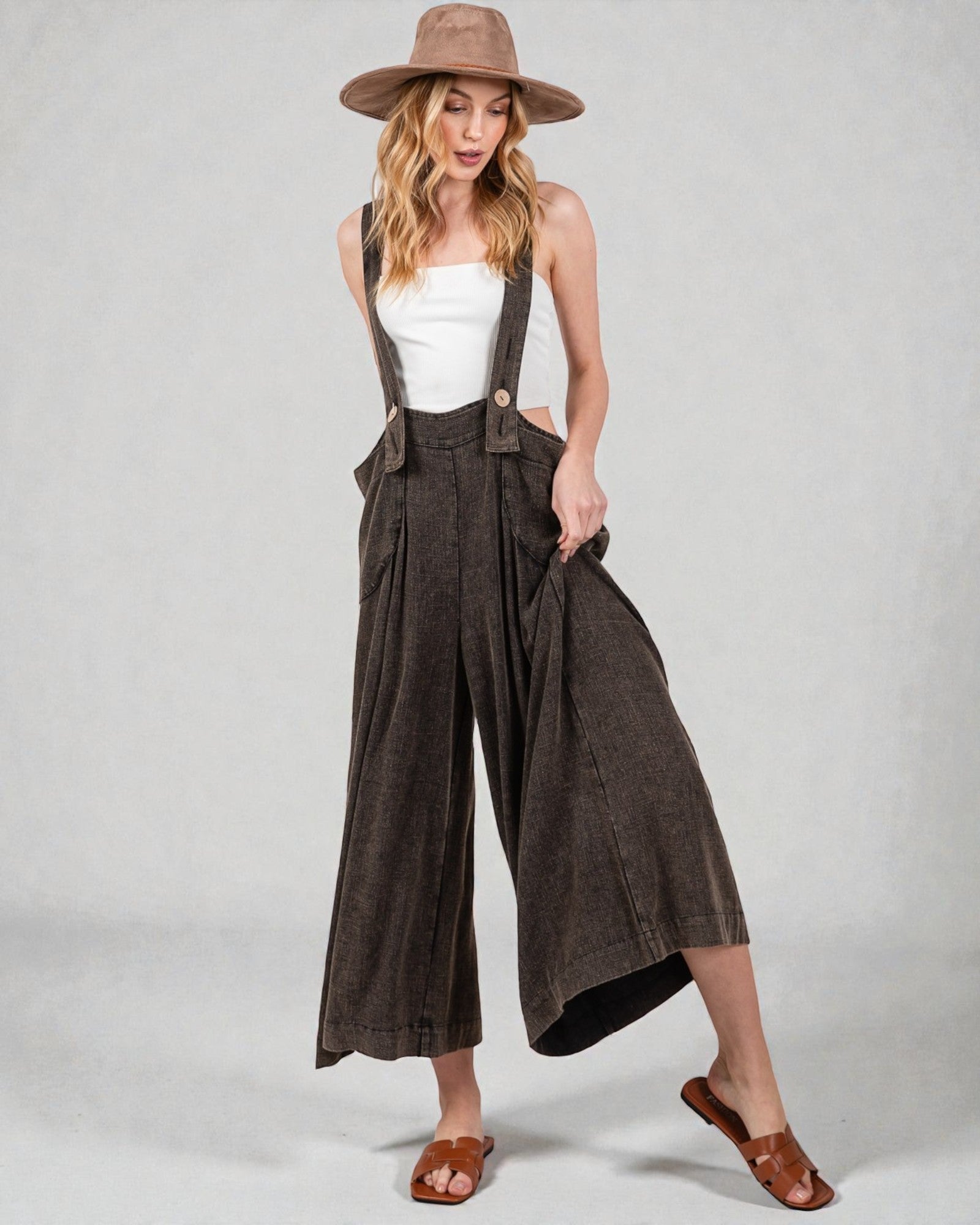 Woman wearing brown wide-leg pants with suspenders and a white top on a plain background