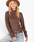 A woman wearing a brown textured knit mock neck sweater, a brown wide-brim hat, and light-wash blue jeans with a brown belt, posed against a solid white background.