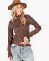 A woman modeling a brown marled knit mock neck sweater, paired with light-wash jeans, a brown belt, and a brown wide-brim hat, against a plain white background.