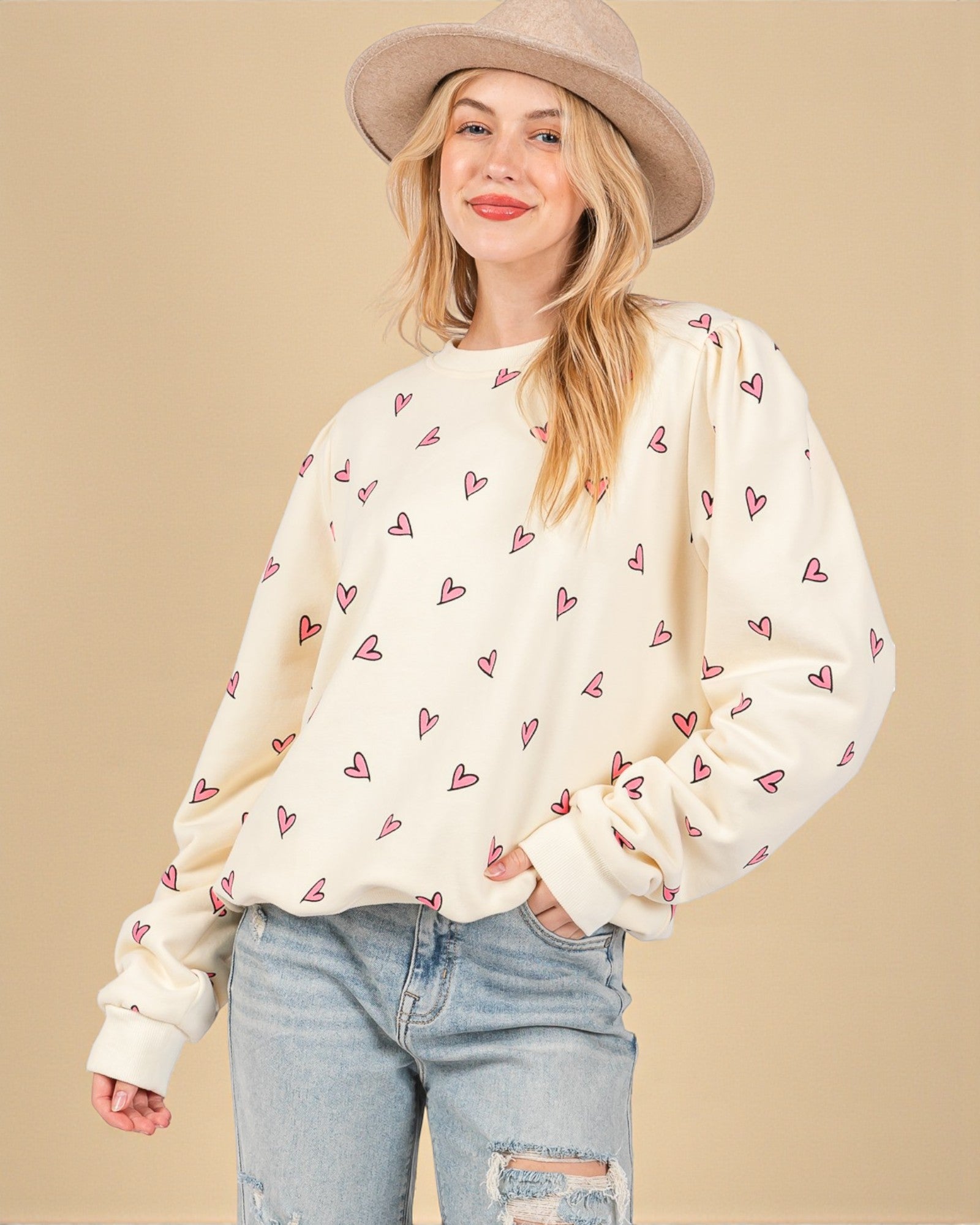 Woman wearing a cream sweater with heart patterns, light blue jeans, and a beige hat on a beige background