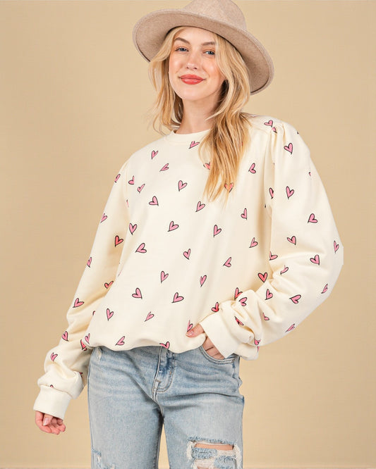 Woman wearing a cream sweater with heart patterns, light blue jeans, and a beige hat on a beige background