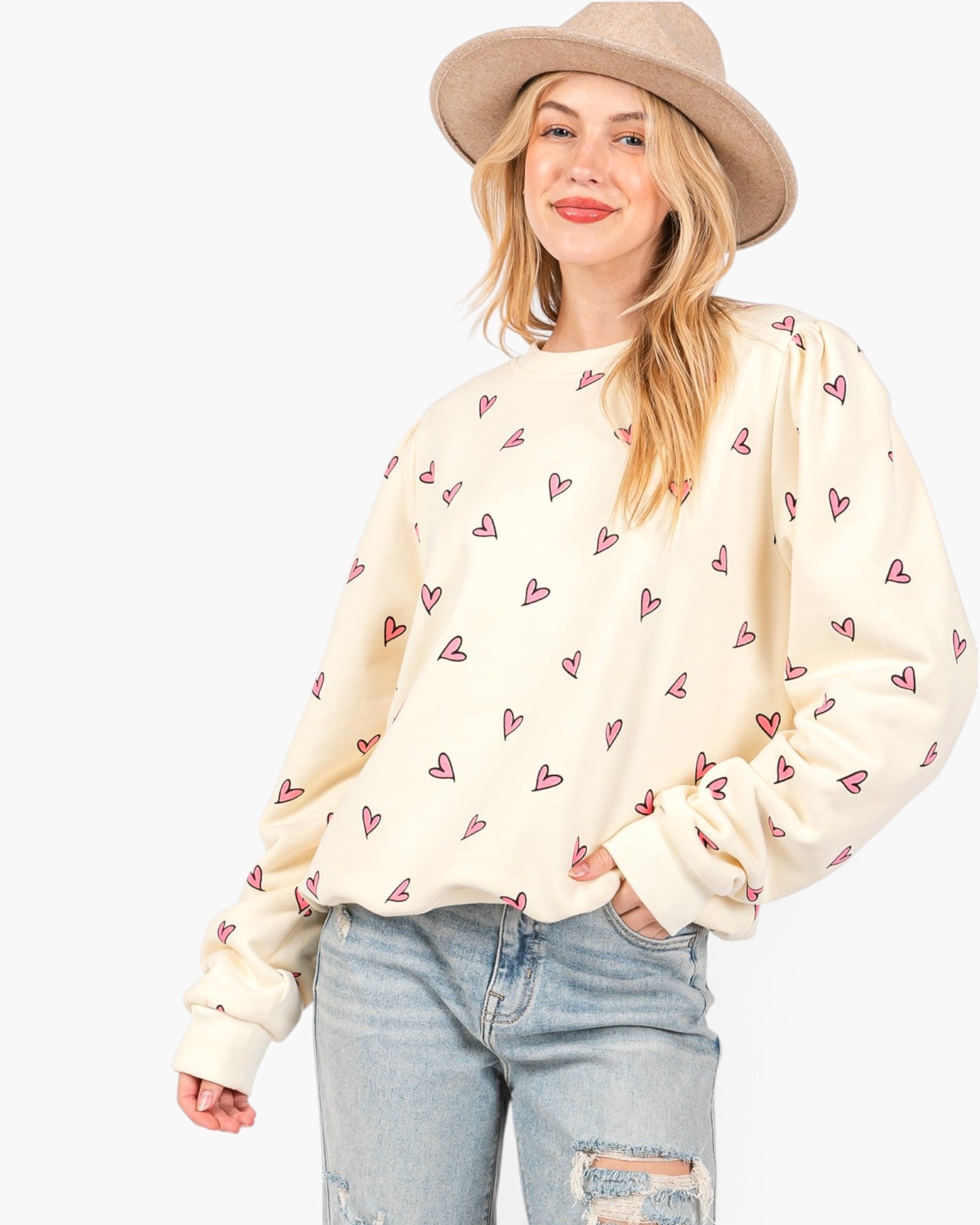 A woman wearing a cream-colored long-sleeve sweatshirt with a repeating pattern of small red hearts, paired with light-wash distressed jeans and a beige wide-brim hat, against a plain white background.