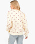 A woman wearing a cream-colored crewneck sweatshirt with a small pink heart pattern, paired with light-wash distressed jeans, against a plain white background.