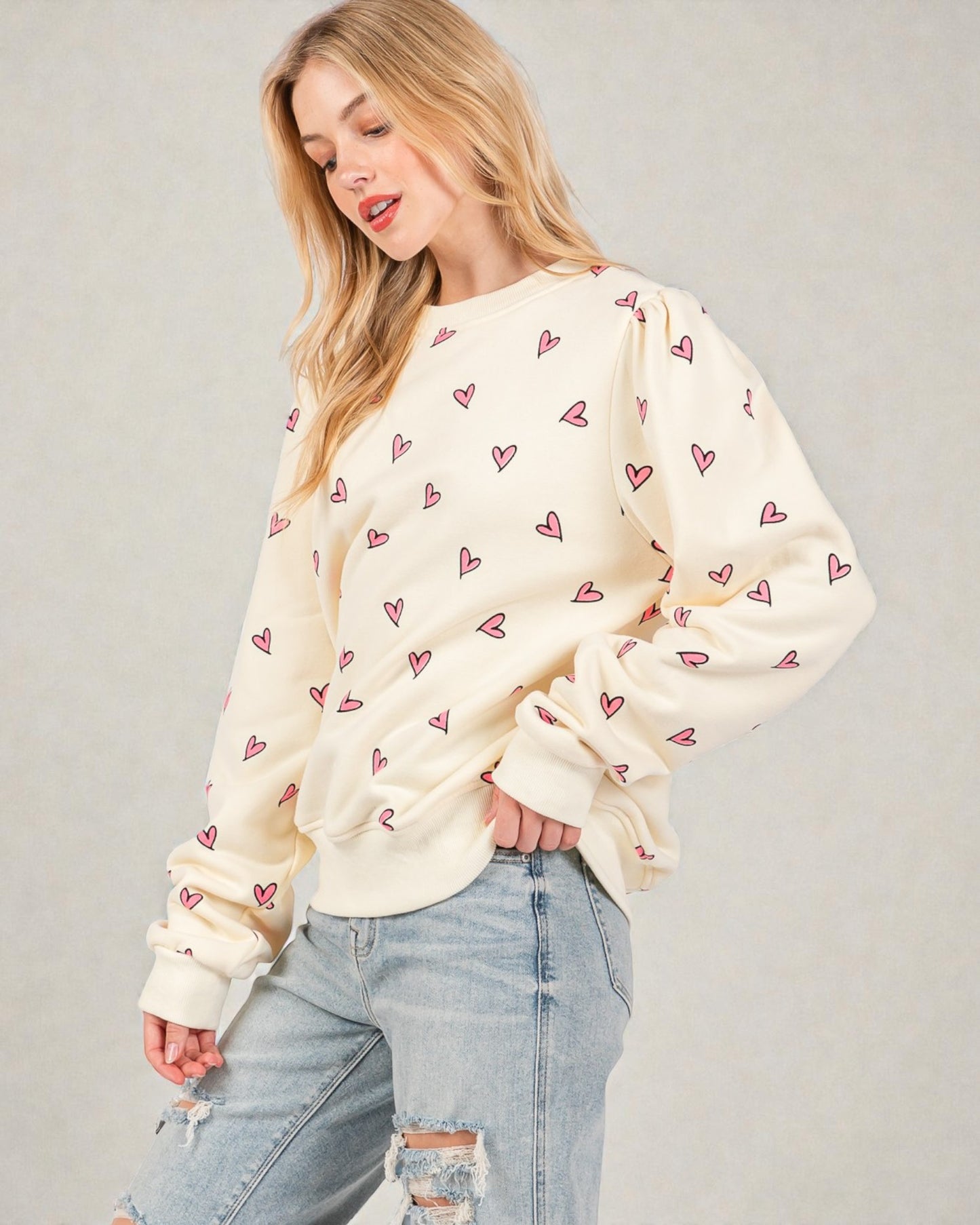 Woman wearing a cream sweatshirt with pink heart patterns and ripped jeans on a plain background