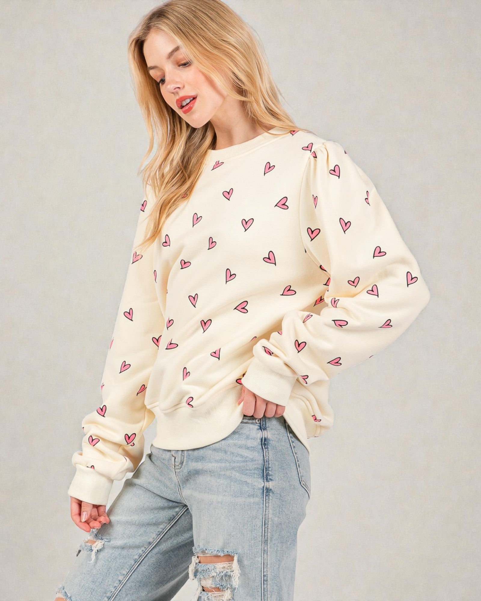 Woman wearing a cream sweatshirt with pink heart patterns and ripped jeans on a plain background