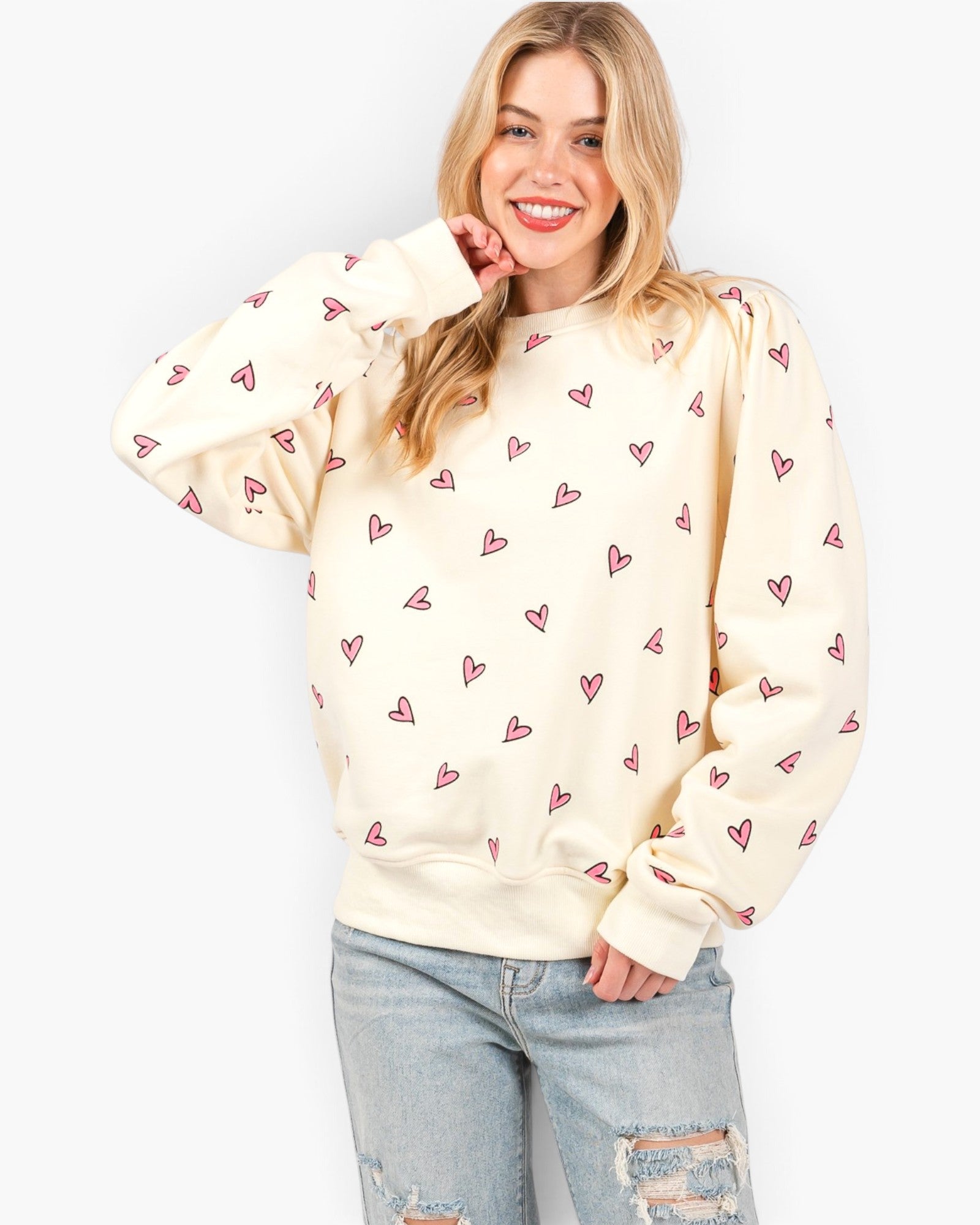 A woman wearing a cream-colored crewneck sweatshirt with a repeating pattern of small pink hearts, paired with light-wash distressed jeans, against a plain white background.