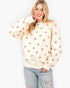 A woman wearing a cream-colored crewneck sweatshirt with a repeating pattern of small pink hearts, paired with light-wash distressed jeans, against a plain white background.