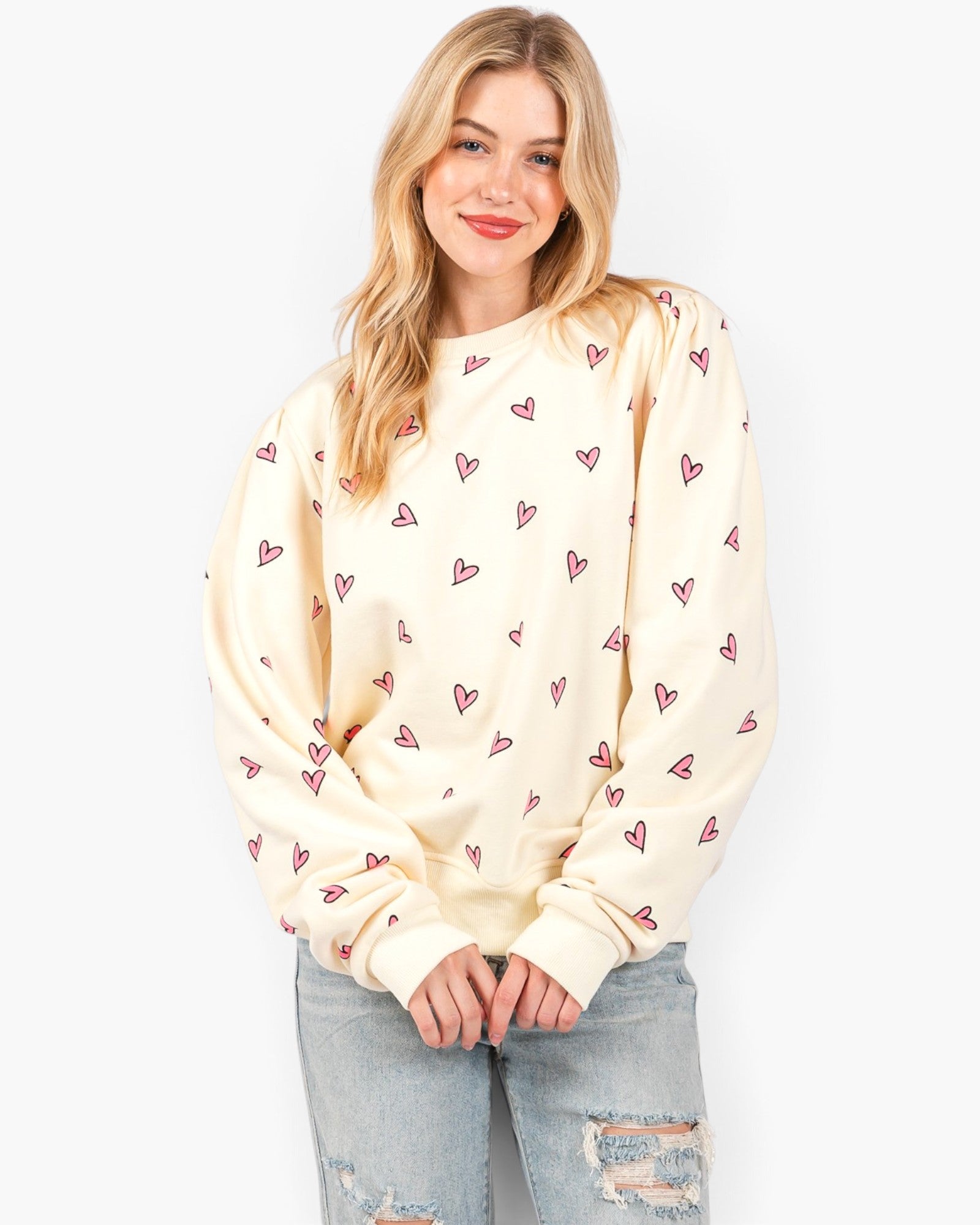 A woman modeling a cream-colored crewneck sweatshirt with a repeating pattern of small pink hearts, paired with light-wash distressed jeans, against a plain white background.