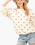 A woman wearing a cream-colored crewneck sweatshirt with a repeating pattern of small pink hearts, paired with light-wash blue jeans and a tan hat, against a plain white background.