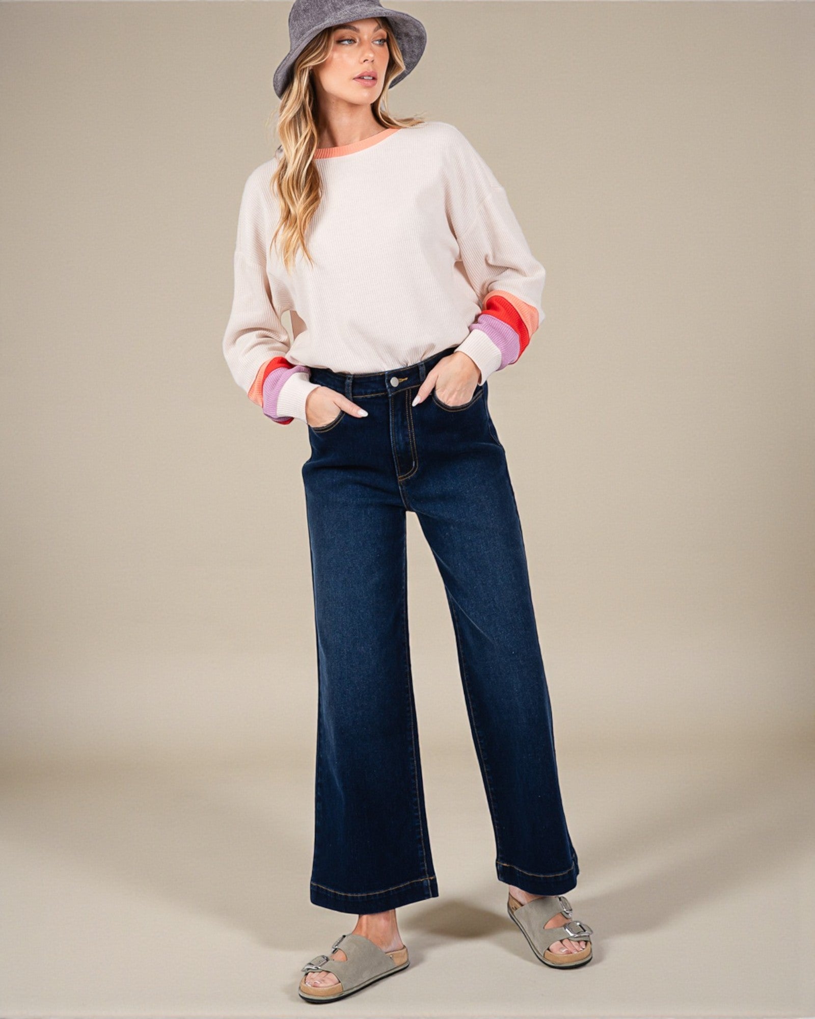 Woman wearing a white sweater with colorful cuffs and hem, blue jeans, and gray sandals on a beige background.