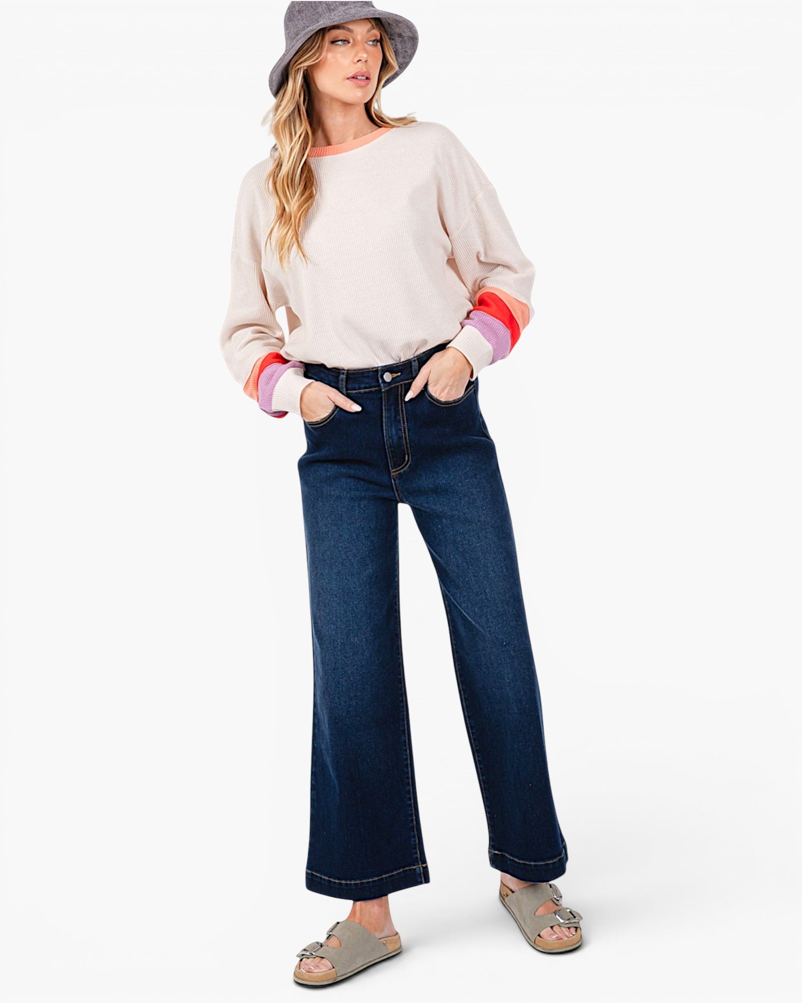 A woman modeling a light beige long-sleeve top with colorful striped cuffs, dark blue wide-leg jeans, a gray bucket hat, and gray slide sandals against a plain white background.