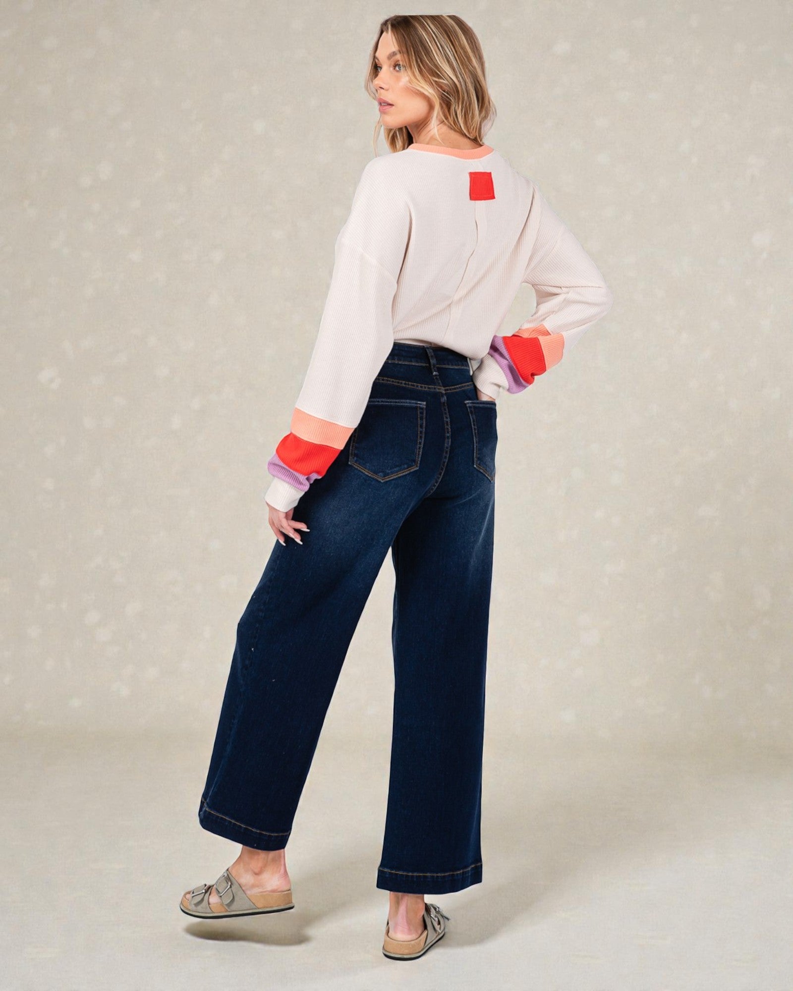 Woman wearing a white sweater with colorful cuffs and blue jeans on a light gray background