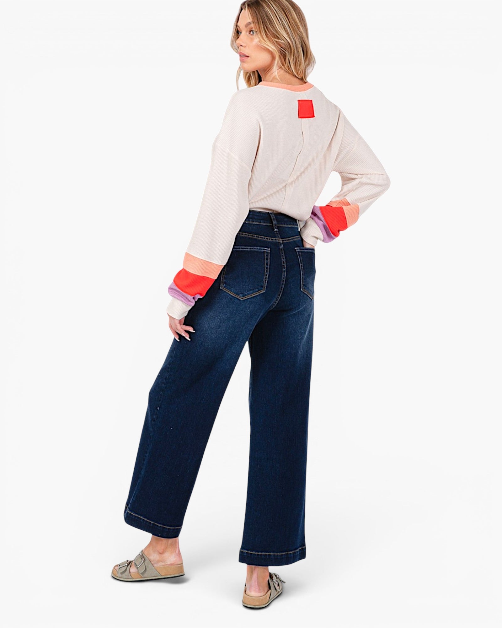A woman modeling a cream long-sleeve top with colorful striped cuffs and dark blue wide-leg jeans, shown from a three-quarters back view against a plain white studio background.
