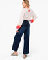 A woman modeling a cream long-sleeve top with colorful striped cuffs and dark blue wide-leg jeans, shown from a three-quarters back view against a plain white studio background.