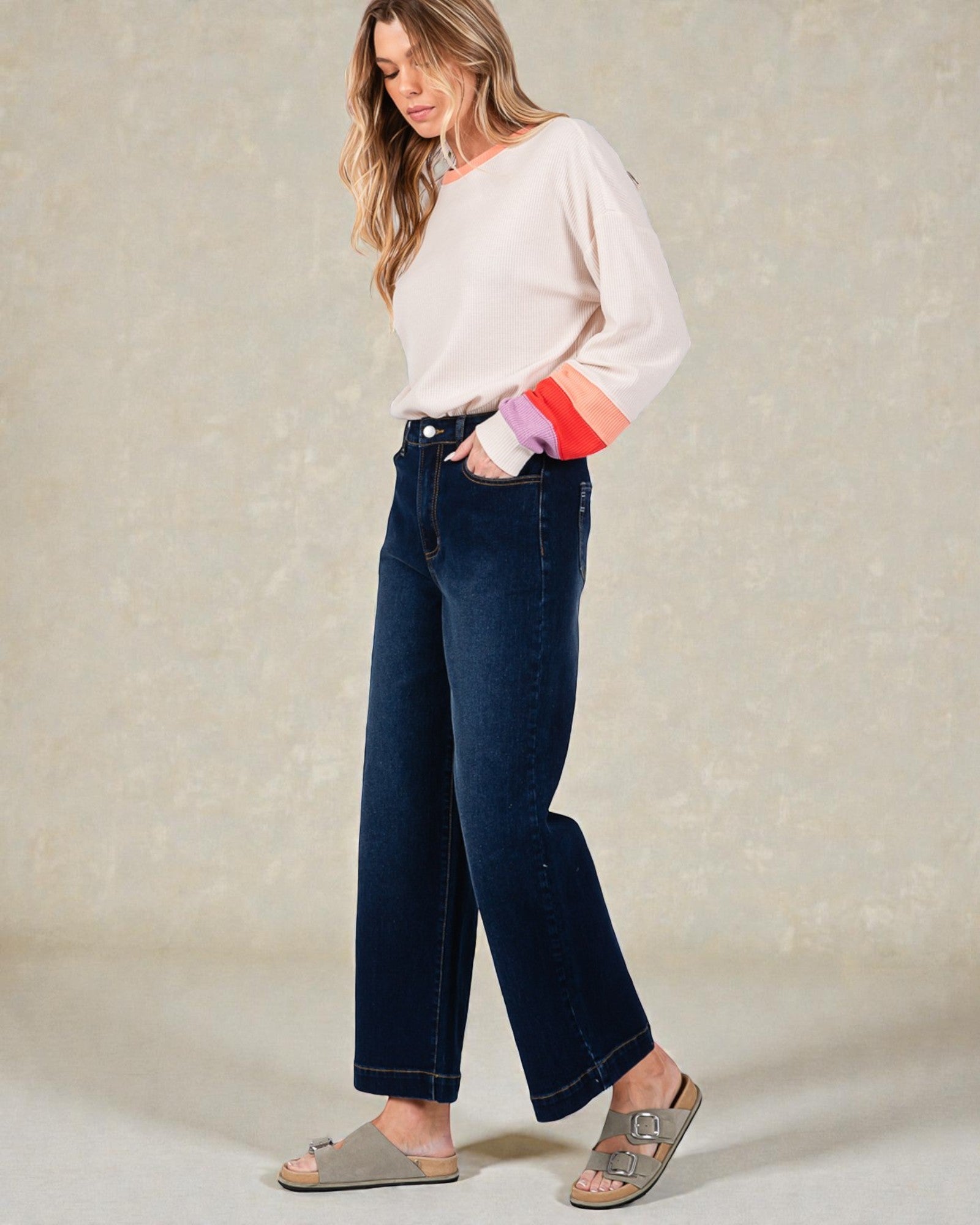 Woman wearing a white sweater with colorful sleeves and dark blue jeans on a neutral background