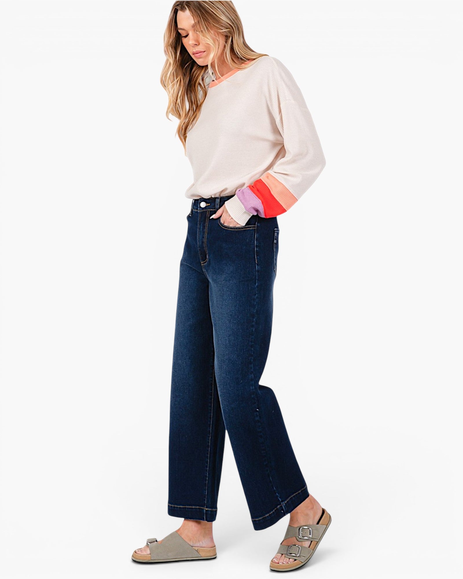 A woman modeling dark blue wide-leg cropped jeans with a cream long-sleeve top and beige slide sandals, standing against a plain white studio background.
