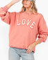 A woman wearing a washed coral pink sweatshirt with 'LOVE' printed across the chest in white varsity letters with red outlines, paired with light blue jeans and a gray cap.
