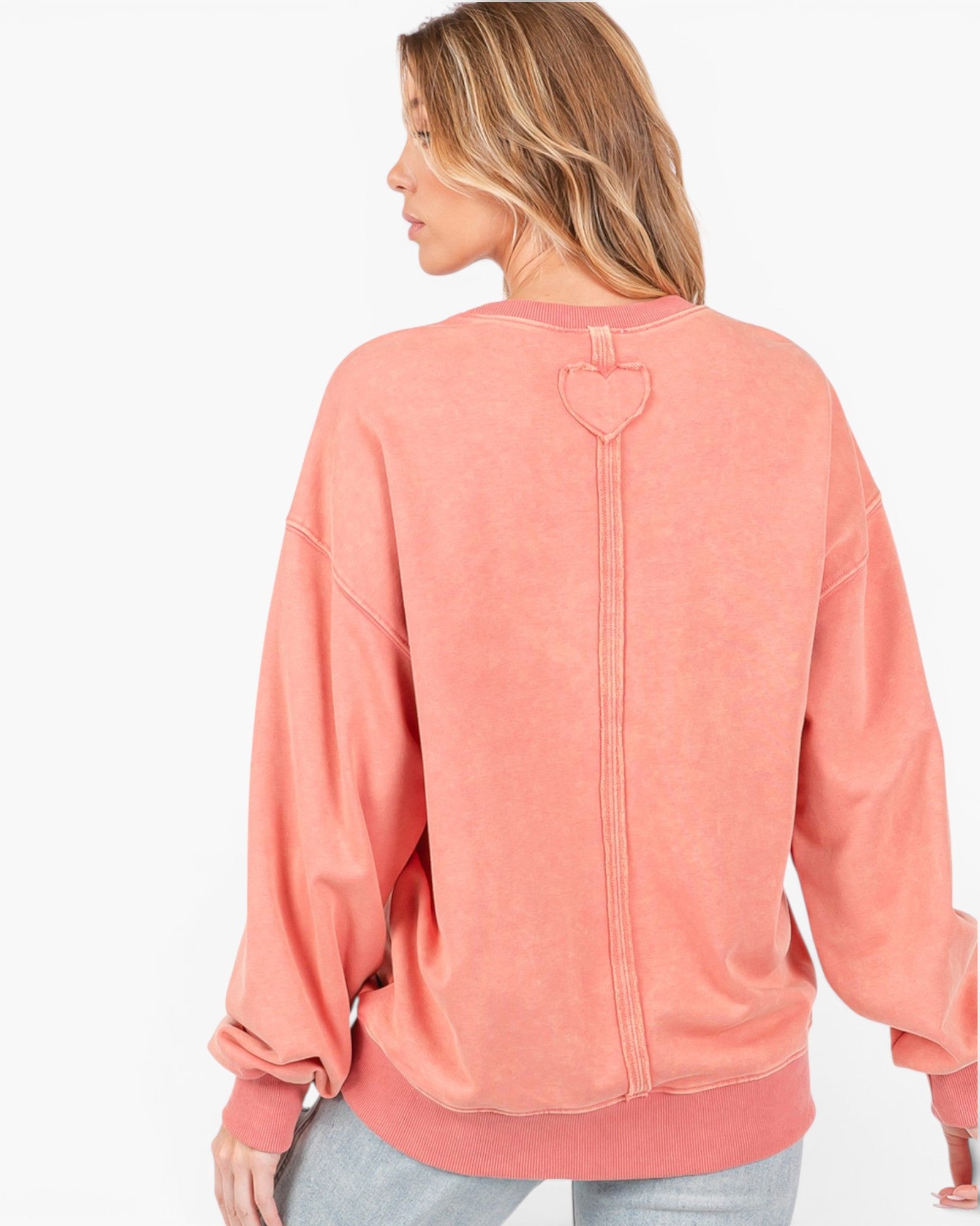 A woman shown from the back wearing a coral pink crewneck sweatshirt with a heart-shaped zipper detail on the back, paired with light gray jeans against a plain white background.