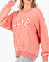A woman wearing a coral pink crewneck sweatshirt with the word 'LOVE' printed in white arched letters across the chest, paired with light blue distressed jeans against a plain white background.