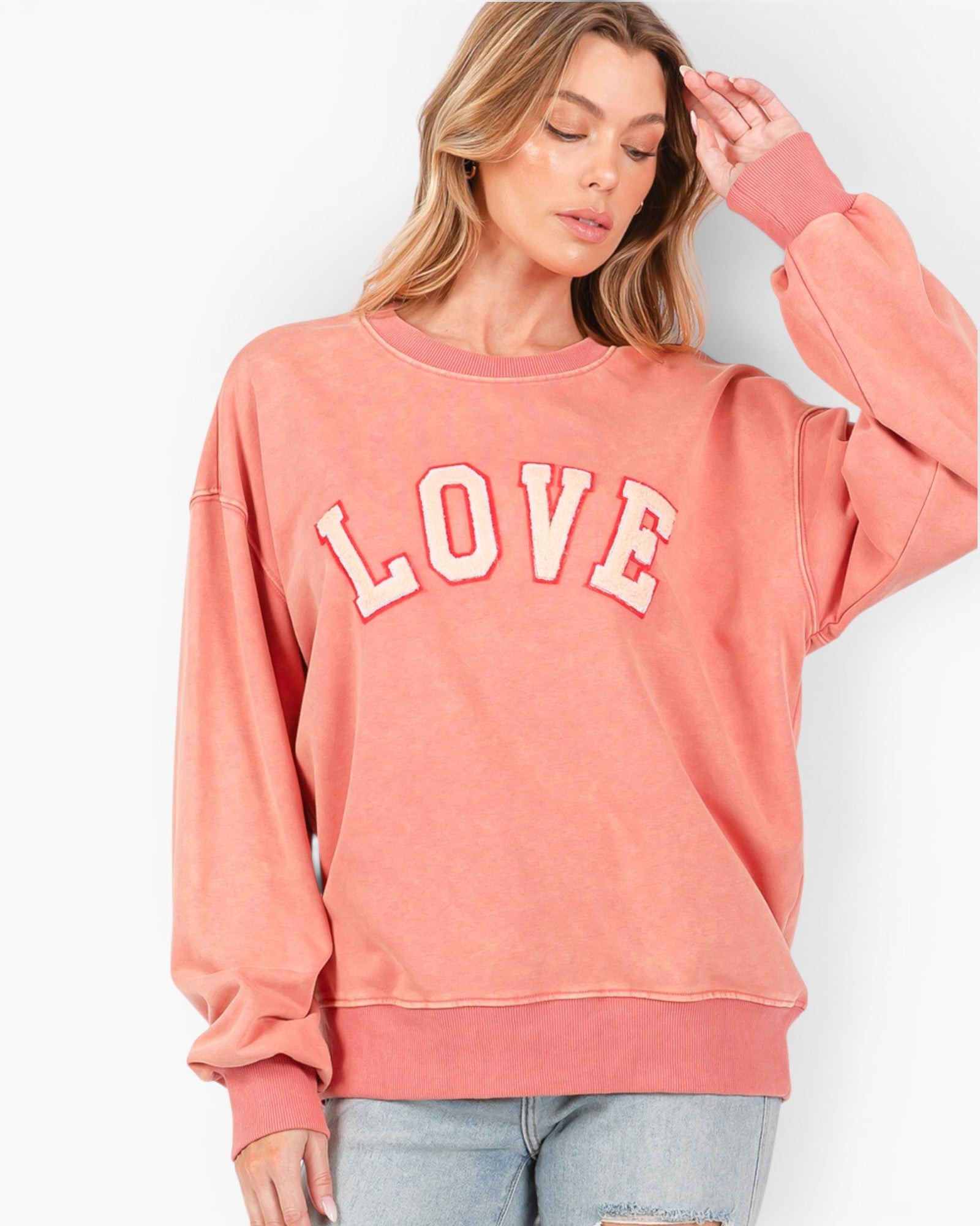 A woman wearing a washed coral-pink crewneck sweatshirt with 'LOVE' printed across the chest in white varsity letters with a pink outline, paired with light-wash jeans against a plain white background.
