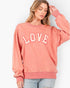 A woman wearing a washed coral-pink crewneck sweatshirt with 'LOVE' printed across the chest in white varsity letters with a pink outline, paired with light-wash jeans against a plain white background.