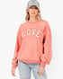 A woman wearing a dusty rose crewneck sweatshirt with 'LOVE' printed in white arched letters across the chest, paired with light-wash distressed jeans and a gray baseball cap, against a plain white background.