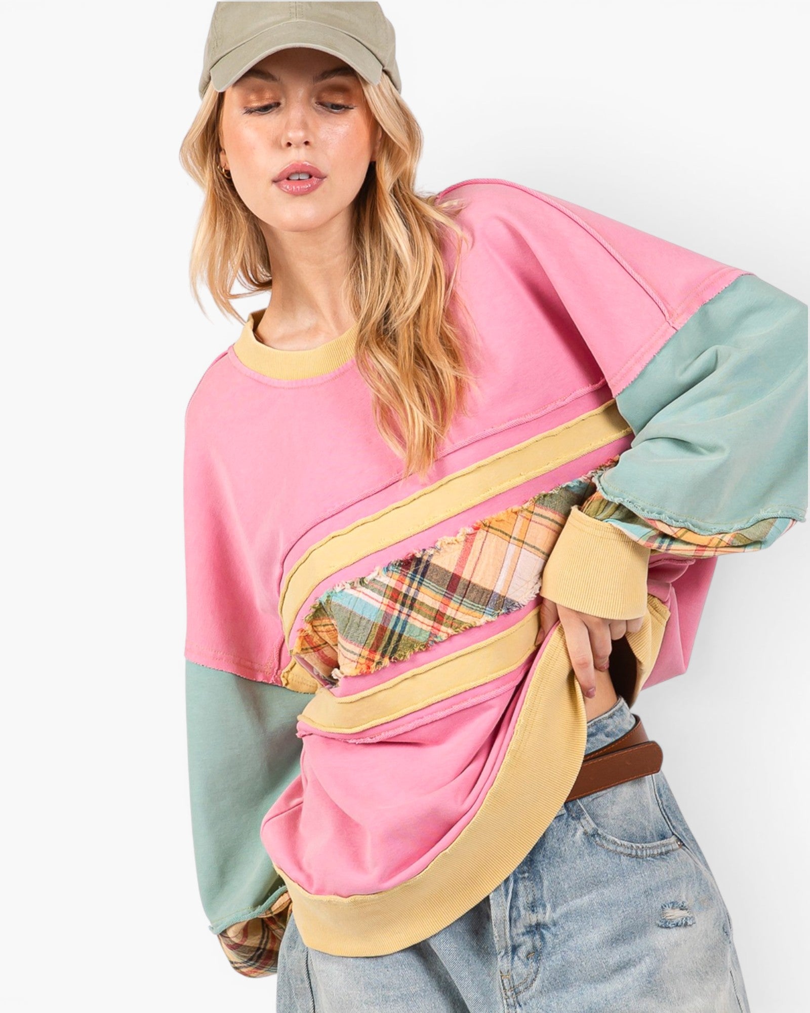 A woman wearing a color-blocked oversized sweatshirt with pink, sage green, and beige panels, paired with light-wash jeans and a khaki cap, against a plain white background.