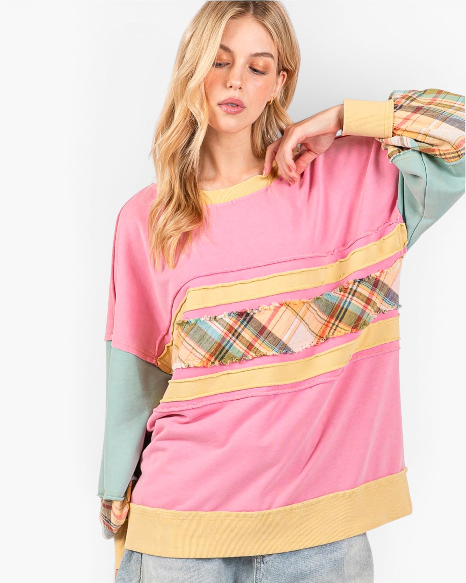 A woman modeling a pink color-blocked sweatshirt with yellow trim, a plaid pattern panel, and long sage green sleeves, set against a plain white background.