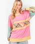 A woman modeling a pink color-blocked sweatshirt with yellow trim, a plaid pattern panel, and long sage green sleeves, set against a plain white background.