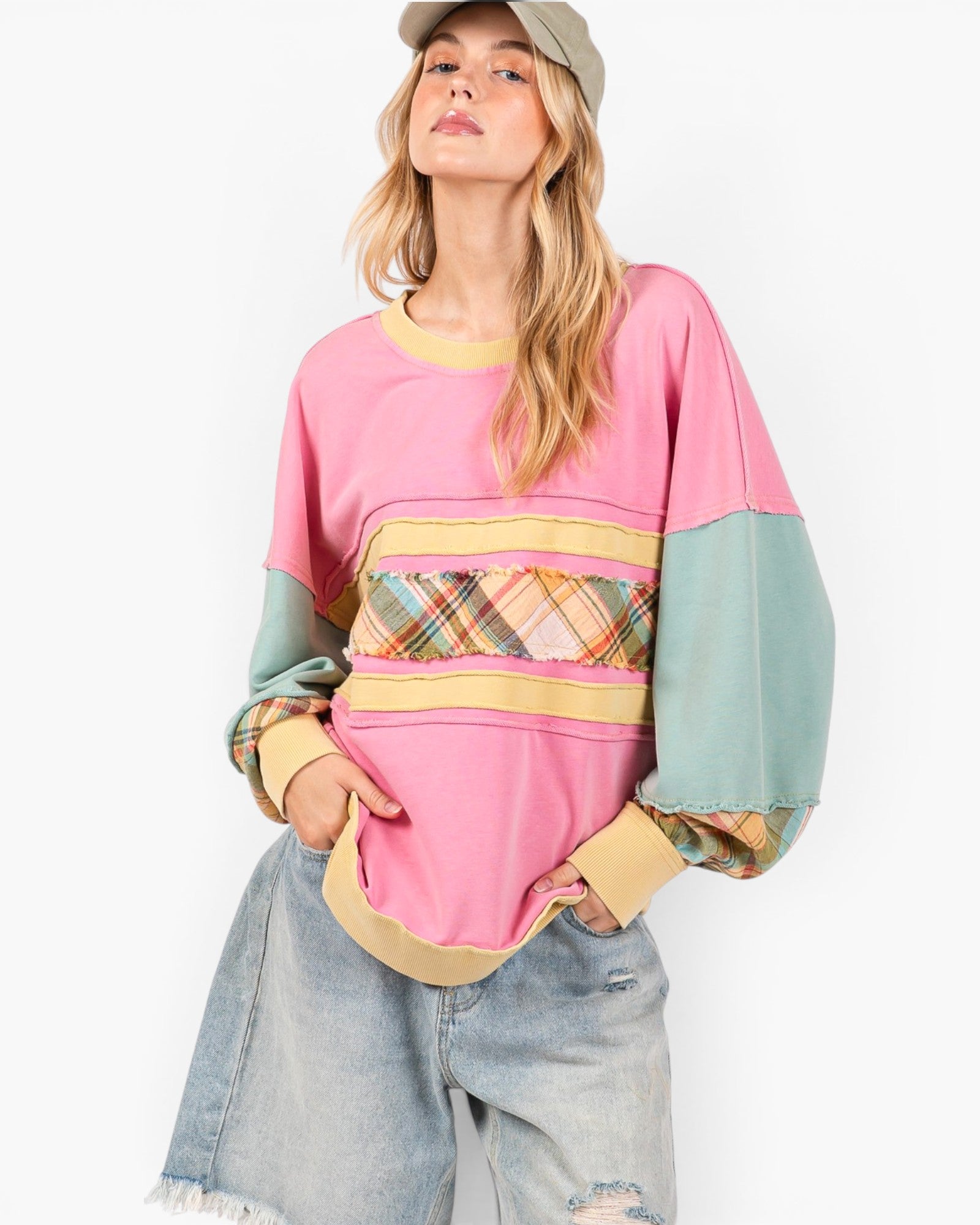 A woman wearing a pastel color-block sweatshirt with pink, mint green, and yellow sections, paired with light-wash denim shorts and a beige cap, against a plain white background.
