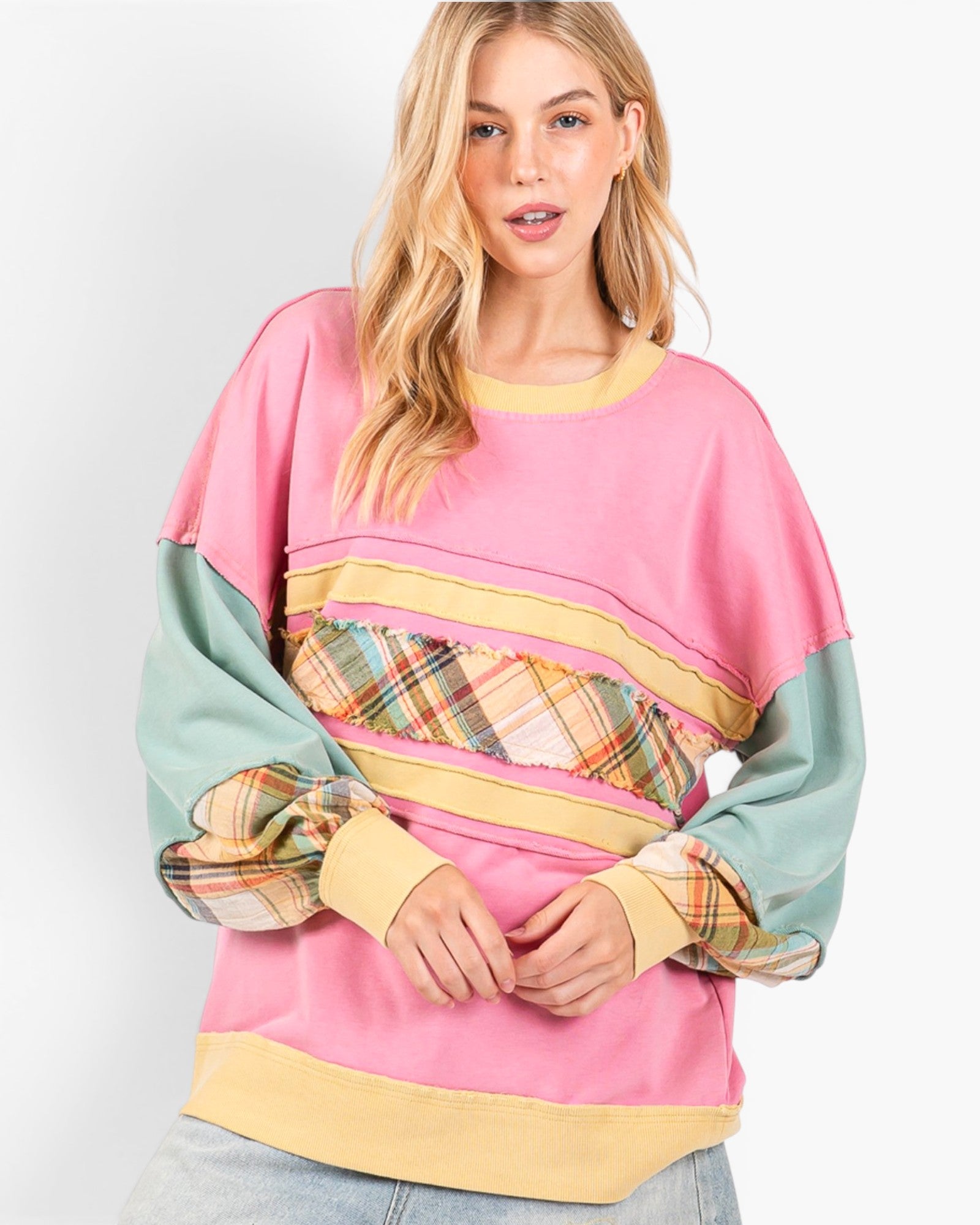 A woman wearing a colorful patchwork sweatshirt with a pink body, sage green sleeves, and horizontal stripes of yellow, plaid, and green, paired with light blue jeans against a light gray studio background.