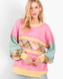 A woman wearing a colorful patchwork sweatshirt with a pink body, sage green sleeves, and horizontal stripes of yellow, plaid, and green, paired with light blue jeans against a light gray studio background.