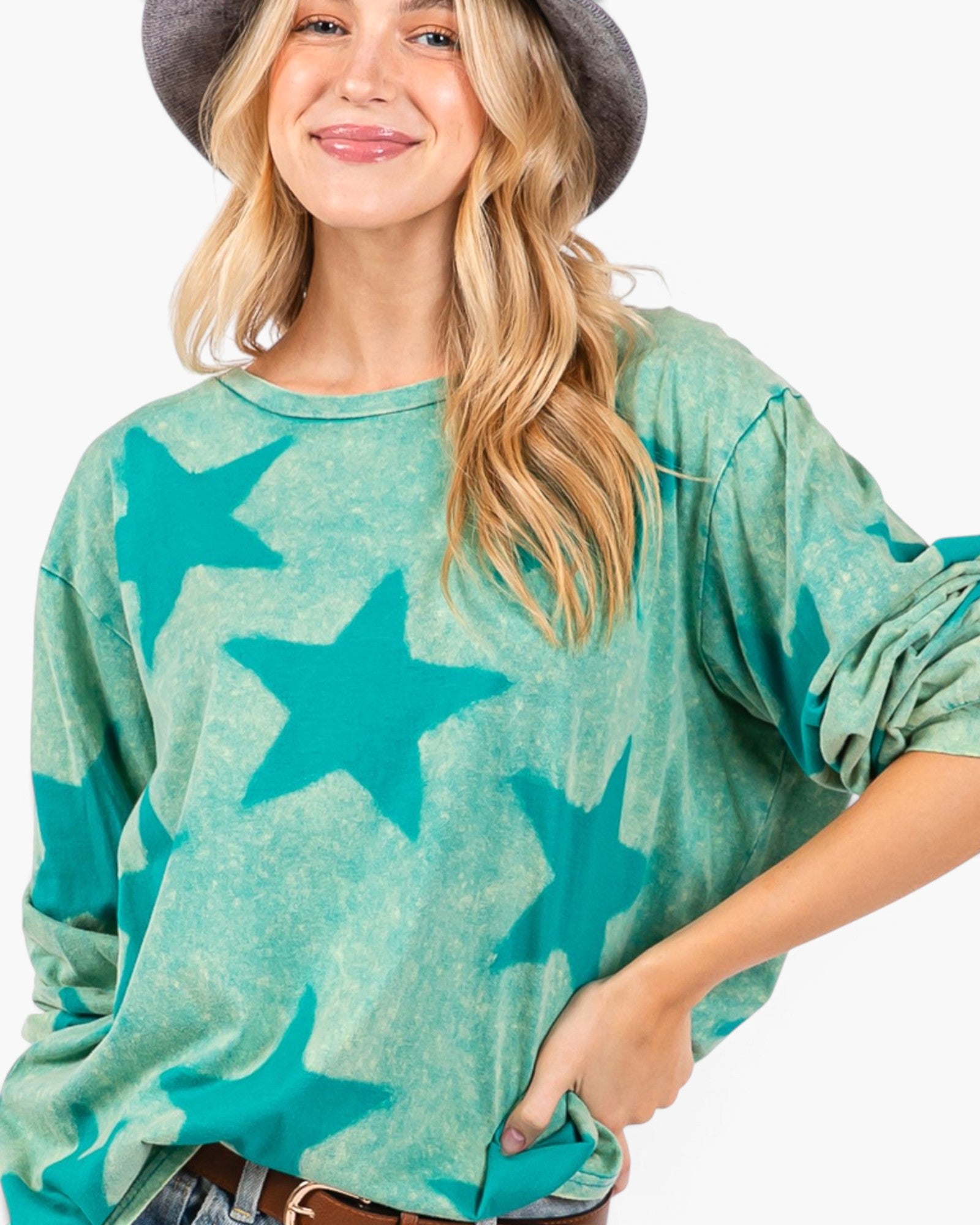 A woman wearing a teal and mint green star-patterned long-sleeve top, styled with a dark gray wide-brim hat and jeans against a plain white background.
