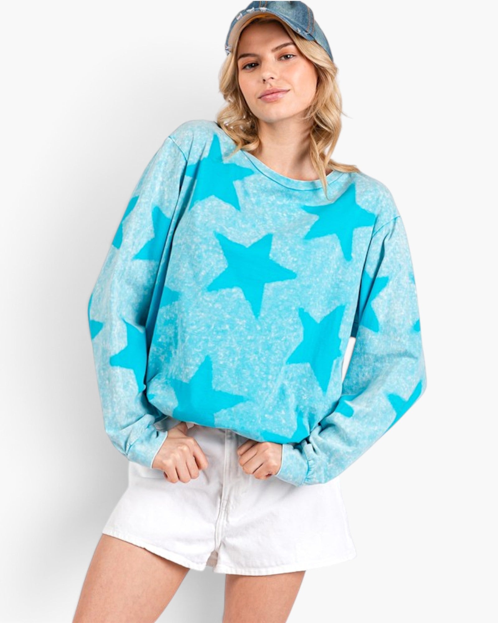A woman wearing a light blue long-sleeve sweatshirt with a darker blue star pattern, paired with white shorts and a denim cap, against a plain white background.