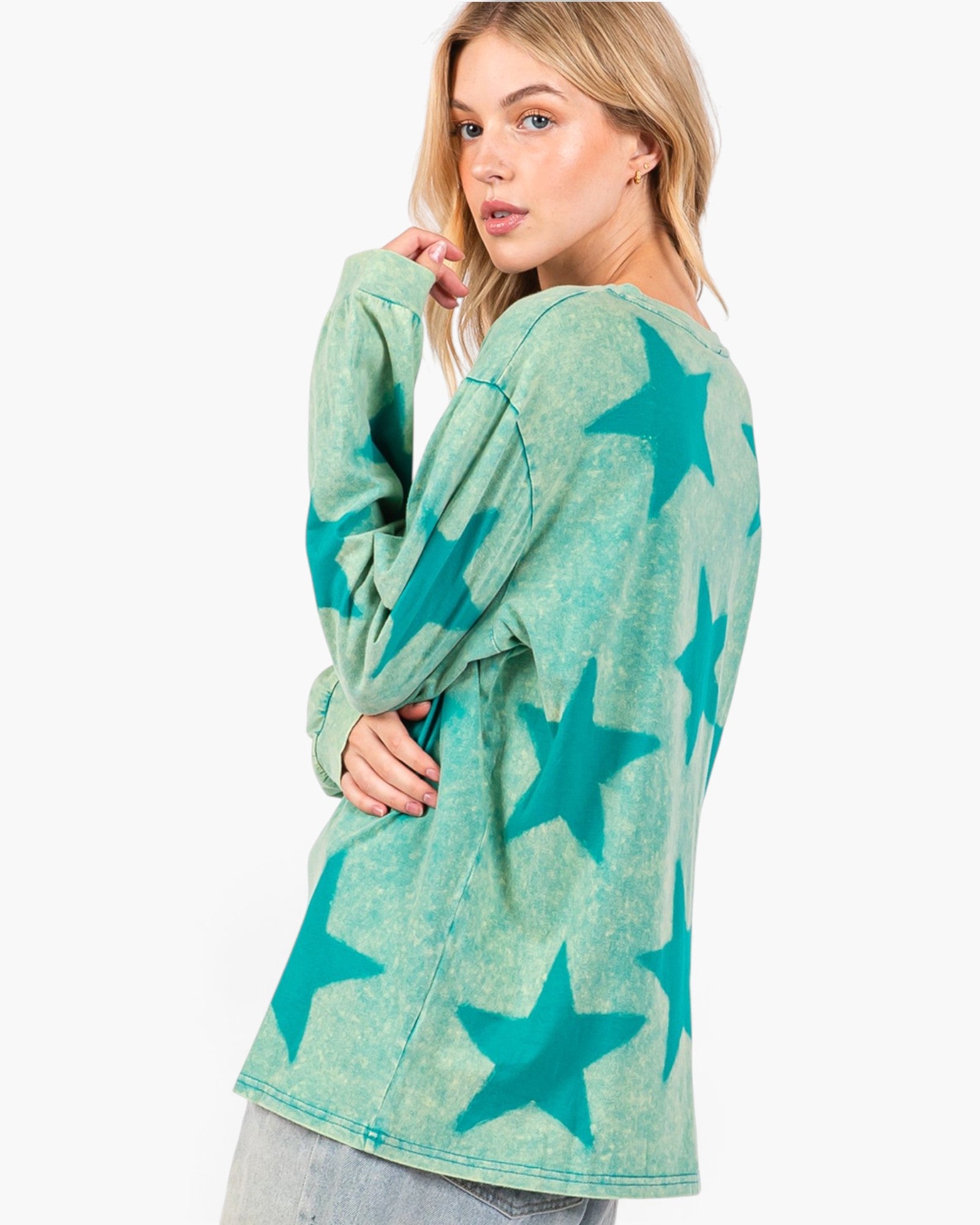 A woman modeling a long-sleeve, oversized t-shirt with a mineral-washed mint green base and a teal star pattern, shown from a three-quarter back view against a plain white background.