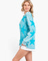 A woman modeling a turquoise blue long-sleeve top with a white tie-dye texture and large star patterns, paired with white shorts, against a plain white background.