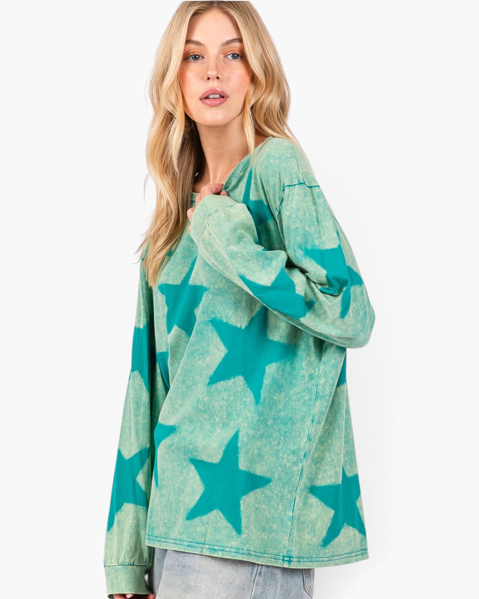 A woman modeling a long-sleeve, oversized crewneck top with a mineral-washed finish and a teal star pattern, paired with light-wash jeans against a plain white studio background.