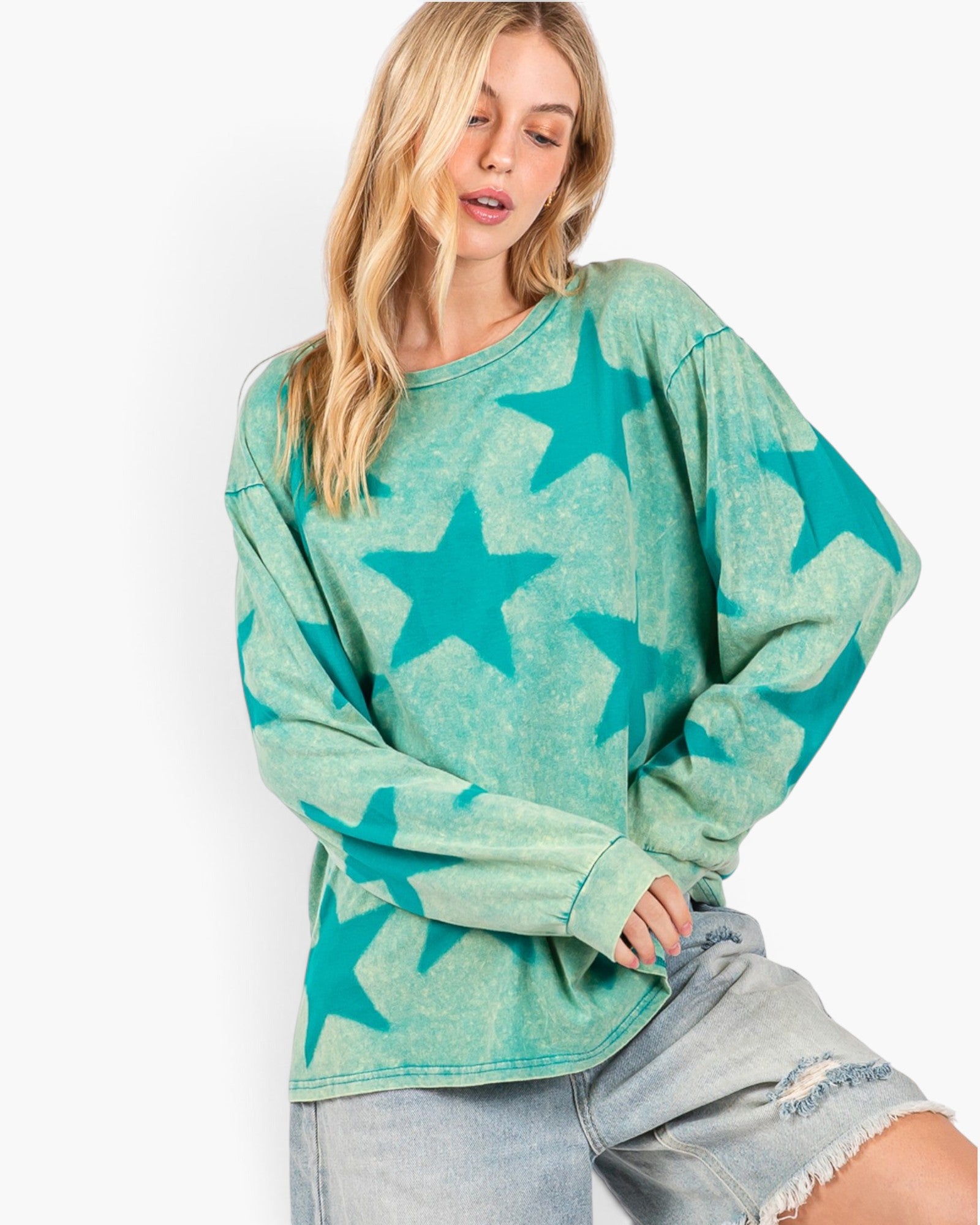 A woman wearing a long-sleeve, oversized crewneck top with a distressed teal and mint green star pattern, paired with light-wash denim shorts, against a plain white background.