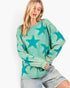 A woman wearing a long-sleeve, oversized crewneck top with a distressed teal and mint green star pattern, paired with light-wash denim shorts, against a plain white background.