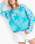A woman wearing a turquoise blue long-sleeve sweatshirt with a white star pattern, paired with white shorts, against a plain white background.