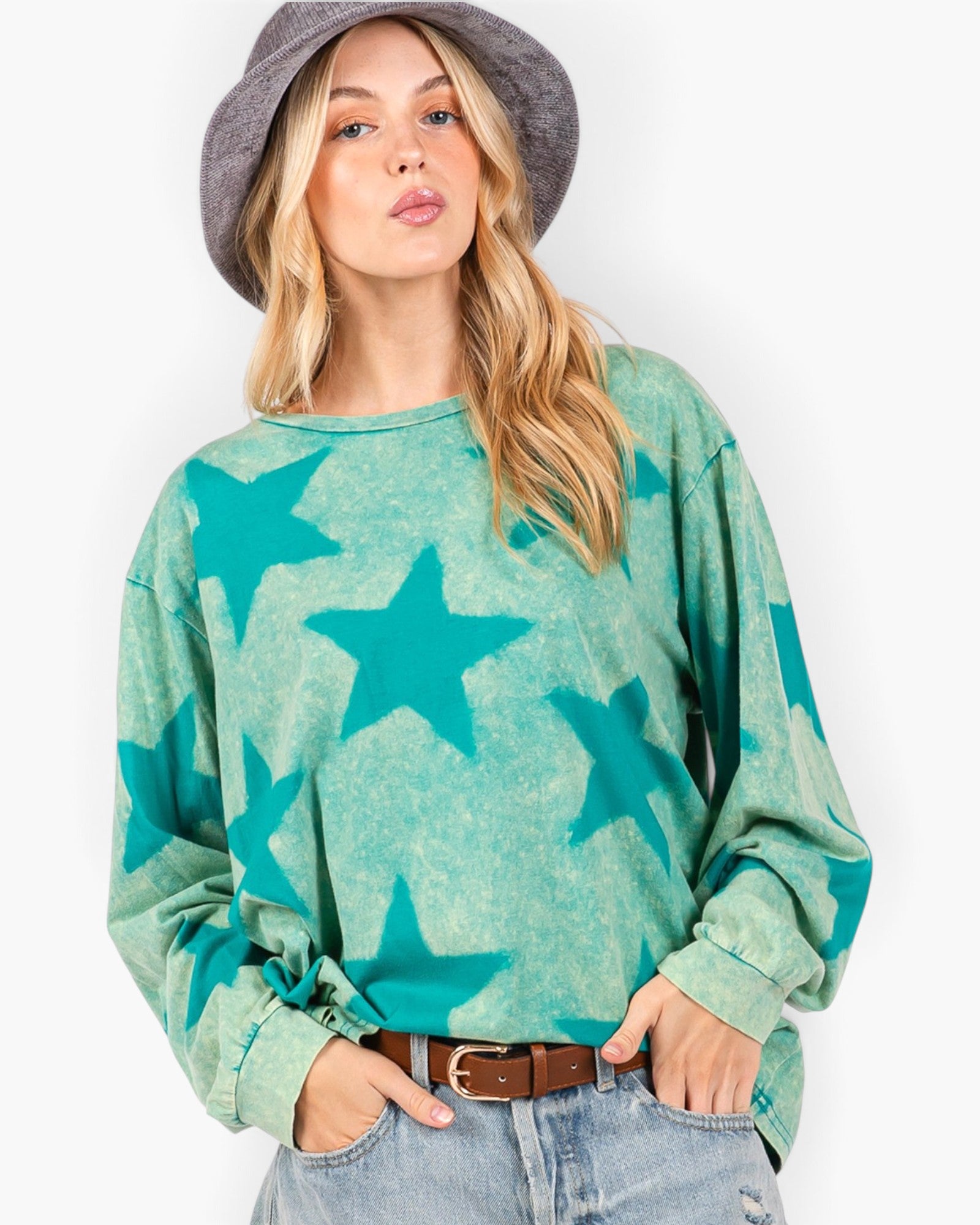 A woman wearing a long-sleeve crewneck sweatshirt with a teal and mint green star pattern, styled with light-wash jeans, a brown belt, and a gray wide-brim hat against a plain white background.