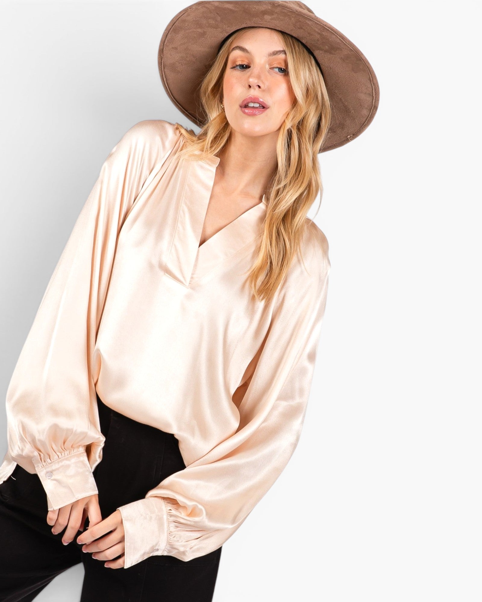 A woman wearing a champagne beige satin long-sleeve blouse with a V-neckline, paired with black pants and a brown wide-brim hat, against a light gray studio background.