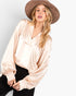 A woman wearing a champagne beige satin long-sleeve blouse with a V-neckline, paired with black pants and a brown wide-brim hat, against a light gray studio background.