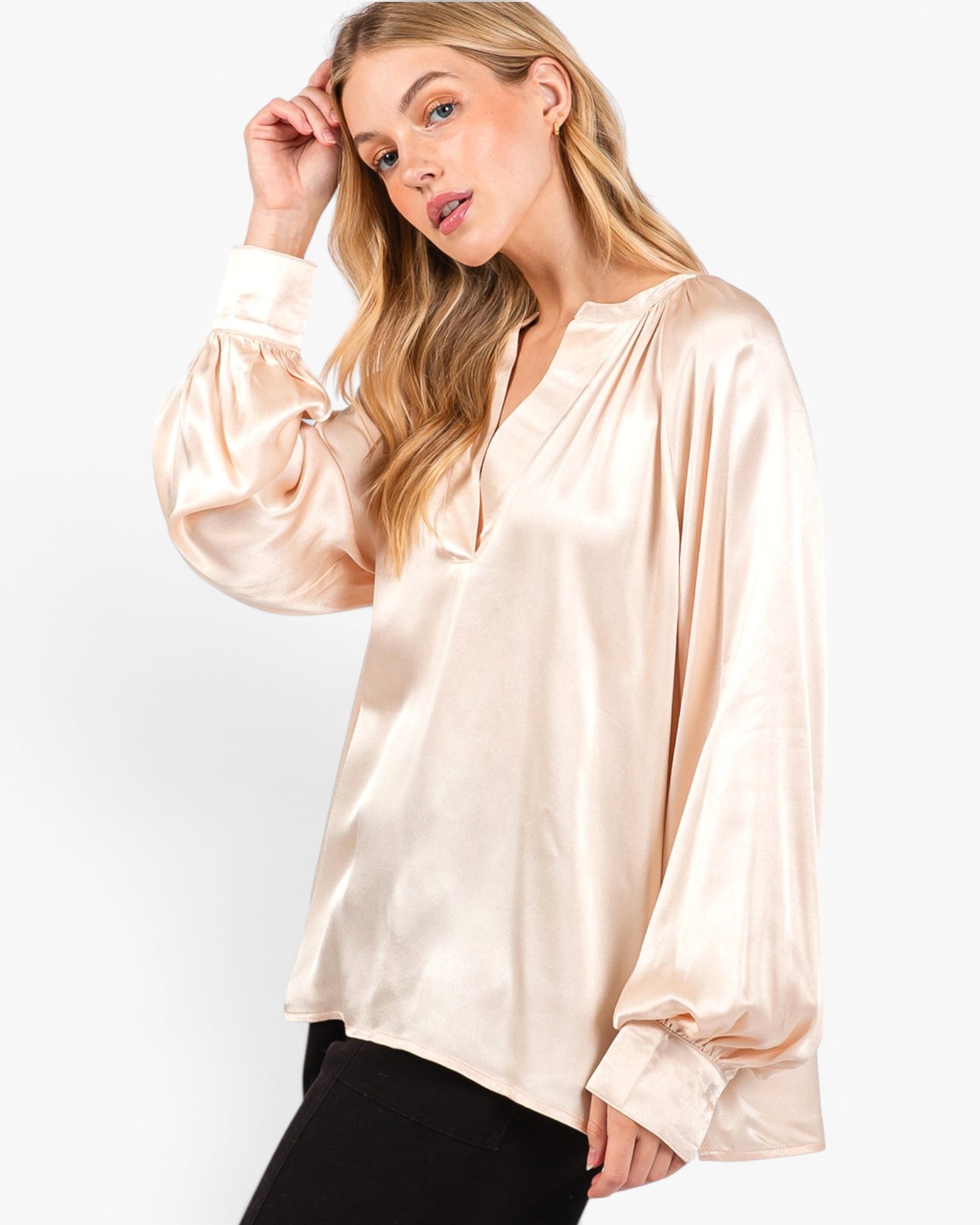 A woman wearing a light beige satin blouse with long balloon sleeves and a split neckline, paired with black pants, against a plain white studio background.
