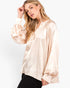 A woman wearing a light beige satin blouse with long balloon sleeves and a split neckline, paired with black pants, against a plain white studio background.
